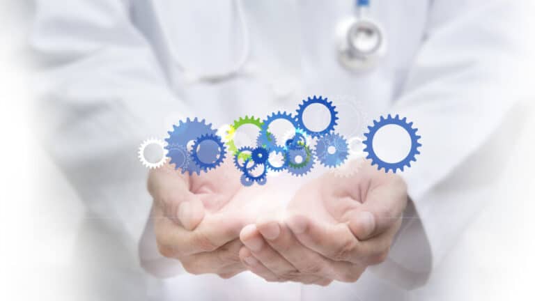 A doctor in a white coat holding out cupped hands, with floating blue and green gears above them, symbolizing mechanism of disease and interplay of biological processes in healthcare. Media credit: © MG / stock.adobe.com