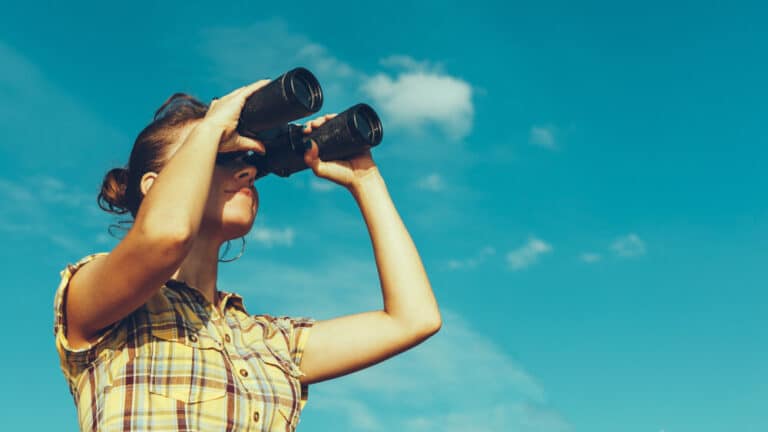 A person in a plaid shirt looking into the distance through binoculars, against a bright, sunny and clear blue sky with a few scattered, white clouds. Suggesting positive outlook for the future and the pursuit of advancements in healthcare. Media credits: © tinyakov / stock.adobe.com