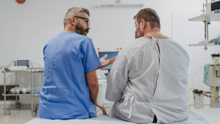 A healthcare professional in blue scrubs sits beside an overweight patient in a hospital gown on a hospital bed, engaged in supportive dialogue. Media credits: © Halfpoint / stock.adobe.com